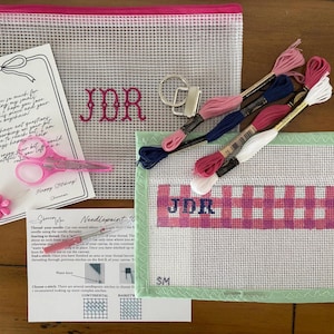 May include: A needlepoint kit featuring a mesh pouch with pink JDR embroidery, a canvas with a pink and blue plaid JDR pattern, four skeins of embroidery floss in pink, blue, and white, a metal keychain, pink scissors, a needle, and instructions.