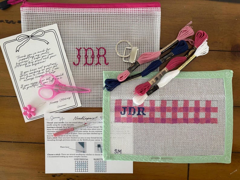 May include: A needlepoint kit featuring a mesh pouch with pink JDR embroidery, a canvas with a pink and blue plaid JDR pattern, four skeins of embroidery floss in pink, blue, and white, a metal keychain, pink scissors, a needle, and instructions.