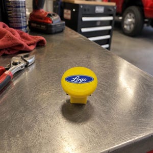 May include: A bright yellow circular knob featuring a central blue oval emblem with the word Logo written in white script. The knob has a smooth, rounded top surface and a square-shaped base, designed for manual operation or adjustment on machinery.