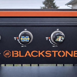May include: A black Blackstone outdoor griddle control panel featuring four circular knobs with Michigan and mascot logos. The panel includes white text markings for heat settings from low to high and an orange brand logo at the bottom centre.