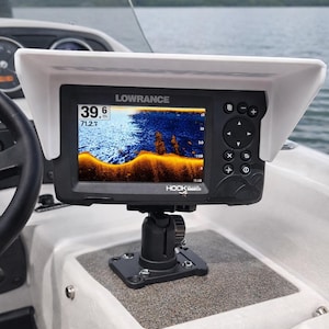 May include: A Lowrance HOOK Reveal fish finder mounted on a boat. The screen displays sonar data with a depth reading of 39.6 feet. The unit is black with a white sunshade, mounted on a black adjustable bracket.