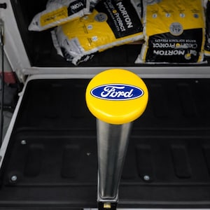 May include: A yellow Ford logo gear shift knob with a blue logo on a silver metal shaft. The background shows bags of water softener pellets.