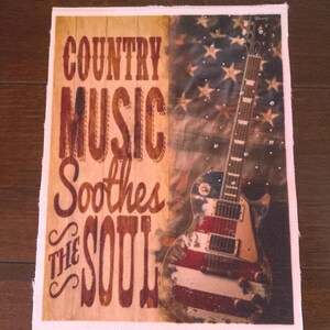 May include: A rectangular fabric panel featuring a vertical wood-grain background on the left with the text "COUNTRY MUSIC Soothes THE SOUL" in brown lettering. The right side displays an electric guitar with an American flag pattern overlay.