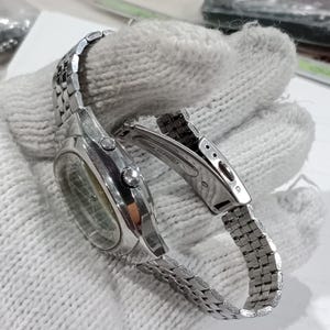 Puede incluir: Un reloj de pulsera de acero inoxidable en tono plateado con caja redonda, brazalete de eslabones met&aacute;licos y cierre desplegable. El lateral de la caja muestra dos botones y una corona central, con el texto stainless steel visible.