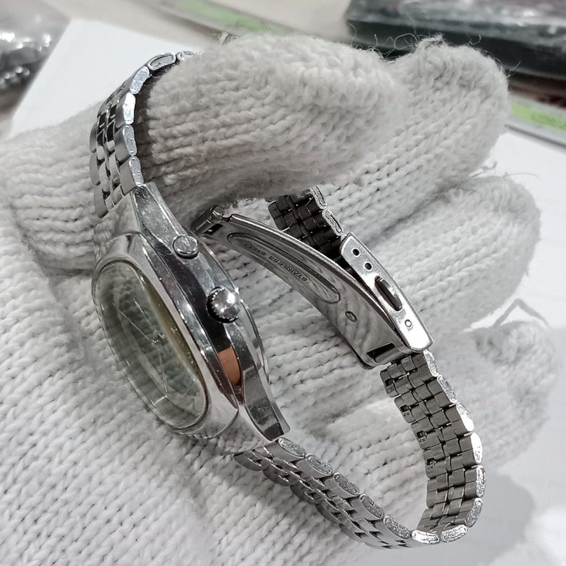 Puede incluir: Un reloj de pulsera de acero inoxidable en tono plateado con caja redonda, brazalete de eslabones met&aacute;licos y cierre desplegable. El lateral de la caja muestra dos botones y una corona central, con el texto stainless steel visible.