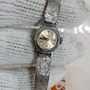 Op de afbeelding: Vintage Rado polshorloge met een zilverkleurige metalen band en een ronde wijzerplaat. De wijzerplaat is lichtcr&egrave;me met zilverkleurige wijzers en markeringen. De band heeft een textuur, geweven ontwerp. Een klassiek accessoire.