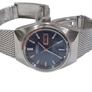 May include: A silver-toned Citizen watch with a mesh band. The watch face is dark blue with silver hands and markers. The day and date are displayed in a small window. The watch is labeled "Citizen Cosmostar" and "Automatic 20000".