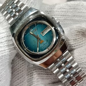 Puede incluir: Un reloj de pulsera autom&aacute;tico Ricoh Crystal en tono plateado con una esfera redonda de color verde azulado y una ventana de d&iacute;a y fecha en la posici&oacute;n de las tres. Incluye una correa de eslabones met&aacute;licos, manecillas plateadas y el texto 21 JEWELS AUTOMATIC.