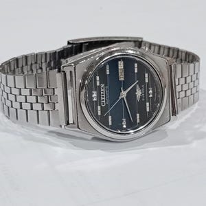 May include: A silver-toned Citizen automatic wristwatch with a dark blue face. The watch features a day and date display, a silver band, and the Citizen logo. The watch is displayed on a white surface.