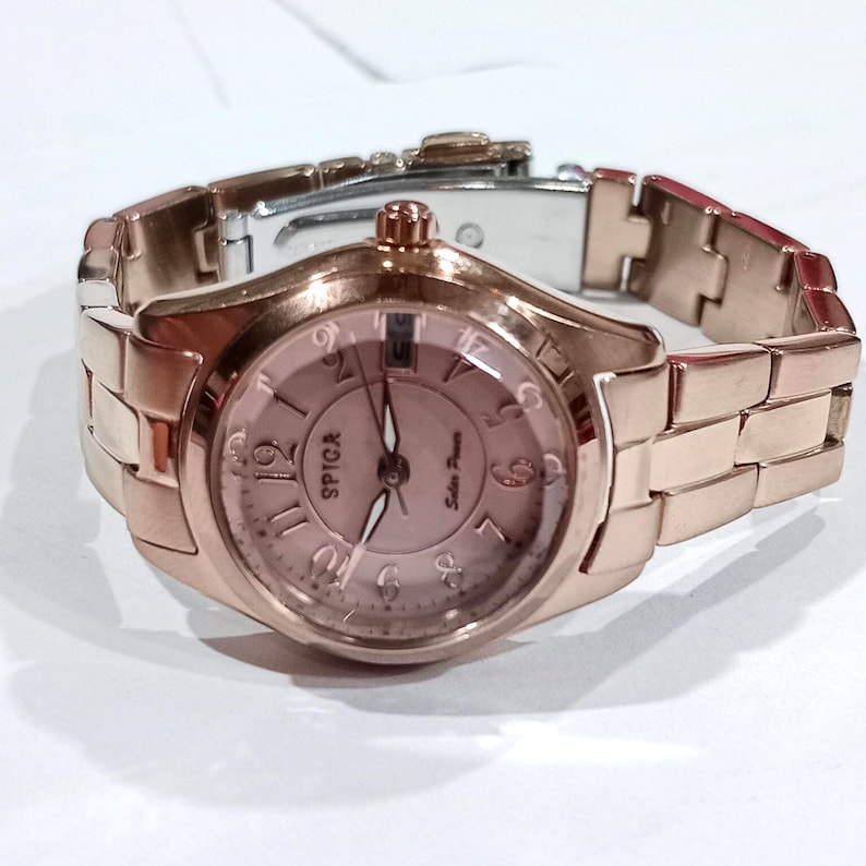 Puede incluir: Un reloj de pulsera de oro rosa con una correa a juego. La esfera del reloj es de color rosa claro con n&uacute;meros y agujas negros. El reloj tiene la palabra "SPICA" impresa en la esfera. La correa est&aacute; hecha de eslabones rectangulares.