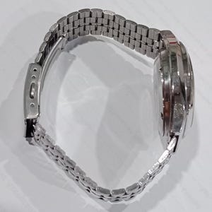 Puede incluir: Un reloj de pulsera plateado con una correa de metal de m&uacute;ltiples eslabones y un cierre desplegable. La caja del reloj es circular con un acabado met&aacute;lico pulido, mostrando una vista lateral del bisel y la estructura de la correa de eslabones.