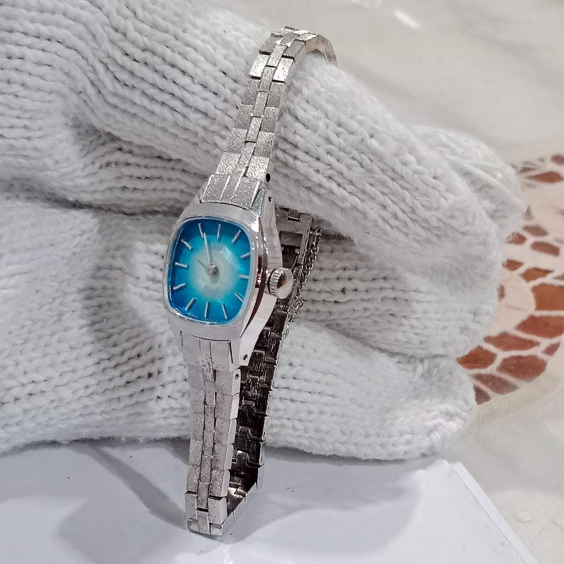 K&ouml;nnte beinhalten: Eine Vintage-Armbanduhr in Silberoptik mit rechteckigem Zifferblatt und blau-wei&szlig;em Farbverlauf. Die Uhr hat ein Gliederarmband und eine kleine Krone. Die Uhr wird auf einer wei&szlig;en, strukturierten Oberfl&auml;che pr&auml;sentiert.