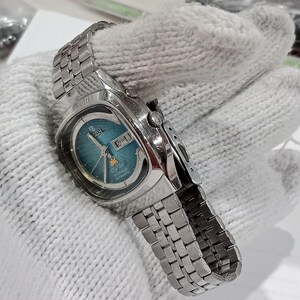 Puede incluir: Un reloj de pulsera autom&aacute;tico Ricoh de tono plateado con una esfera redonda de color verde azulado con efecto rayos de sol, una ventana de d&iacute;a y fecha en la posici&oacute;n de las tres, un peque&ntilde;o emblema de estrella dorada y una correa de acero.