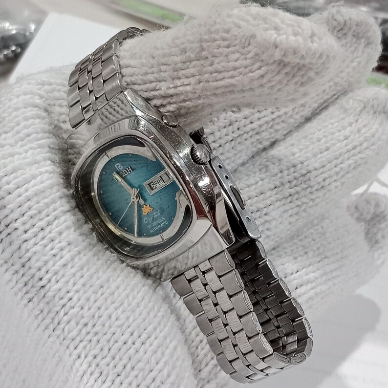 Puede incluir: Un reloj de pulsera autom&aacute;tico Ricoh de tono plateado con una esfera redonda de color verde azulado con efecto rayos de sol, una ventana de d&iacute;a y fecha en la posici&oacute;n de las tres, un peque&ntilde;o emblema de estrella dorada y una correa de acero.