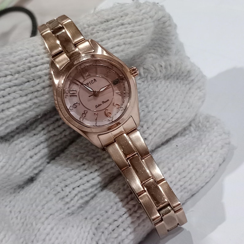 Puede incluir: Un reloj de pulsera de oro rosa con esfera redonda y brazalete de eslabones a juego. La esfera del reloj tiene un fondo rosa claro con el nombre de la marca "PIGA" y las palabras "Solar Power" impresas. El reloj se muestra sobre una tela gris.