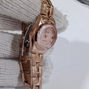 Puede incluir: Reloj de pulsera de color oro rosa con esfera rosa y visualizaci&oacute;n de la fecha. El reloj presenta un brazalete de eslabones y una esfera redonda con la marca "SVICA". Un accesorio cl&aacute;sico.
