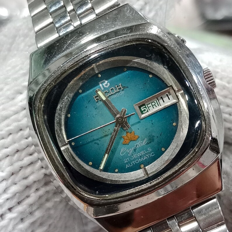 Puede incluir: Un reloj de pulsera autom&aacute;tico Ricoh de tono plateado con esfera de color verde azulado met&aacute;lico, que presenta una ventana de d&iacute;a y fecha en la posici&oacute;n de las tres, un peque&ntilde;o emblema de hoja dorada y una correa de eslabones de acero inoxidable.