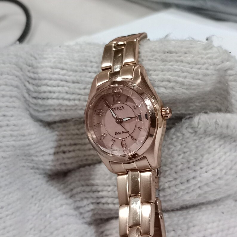 Puede incluir: Un reloj Spica de oro rosa con esfera rosa y correa a juego. El reloj tiene una esfera redonda con un dise&ntilde;o similar a un cristal y una peque&ntilde;a corona. El reloj se muestra sobre una tela blanca.