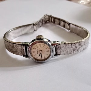 Op de afbeelding: Een vintage Rado polshorloge met een zilverkleurige metalen band en een ronde, cr&egrave;mekleurige wijzerplaat. Het horloge heeft een getextureerde band en een kleine, sierlijke kroon. De wijzerplaat heeft zwarte uurmarkeringen en wijzers.