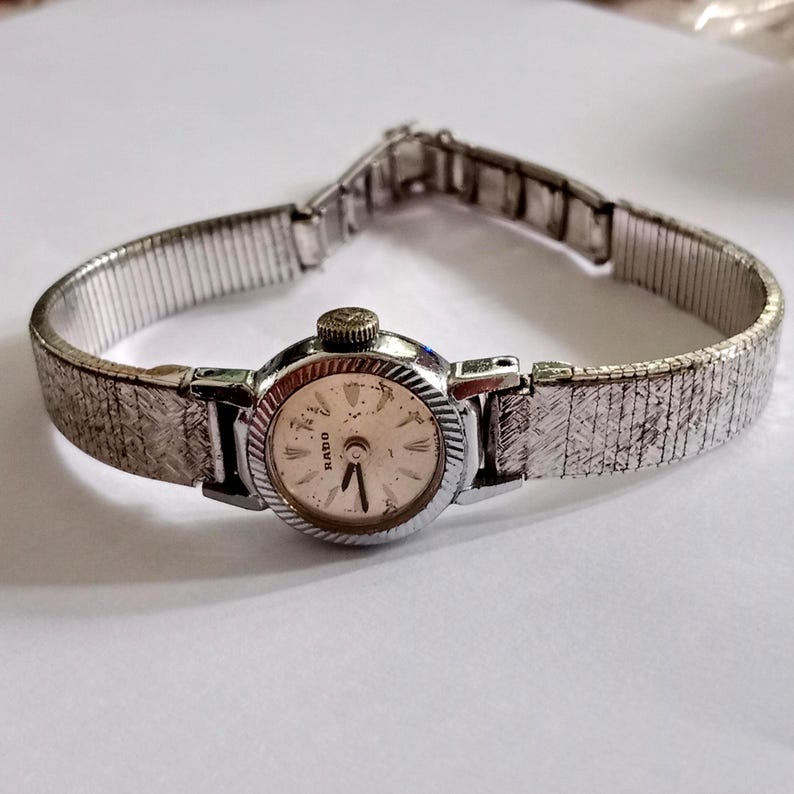 Op de afbeelding: Een vintage Rado polshorloge met een zilverkleurige metalen band en een ronde, cr&egrave;mekleurige wijzerplaat. Het horloge heeft een getextureerde band en een kleine, sierlijke kroon. De wijzerplaat heeft zwarte uurmarkeringen en wijzers.