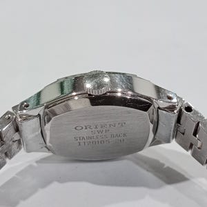 K&ouml;nnte beinhalten: Nahaufnahme einer silberfarbenen Armbanduhr. Die Uhr hat einen Edelstahlboden mit dem Text "ORIENT SWP STAINLESS BACK I120105-20". Das Armband der Uhr ist ebenfalls silberfarben und besteht aus Metallgliedern.