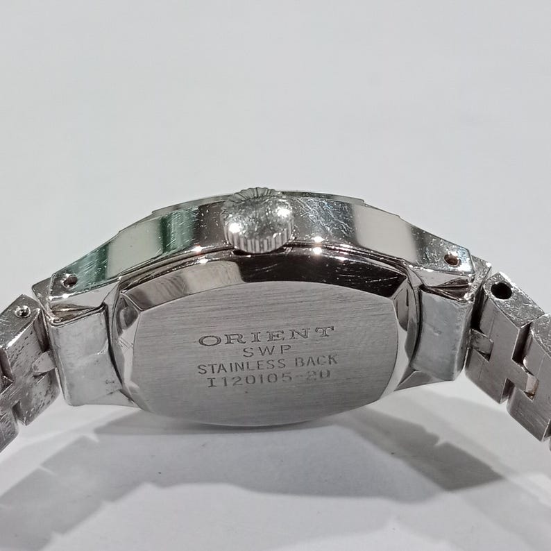 K&ouml;nnte beinhalten: Nahaufnahme einer silberfarbenen Armbanduhr. Die Uhr hat einen Edelstahlboden mit dem Text "ORIENT SWP STAINLESS BACK I120105-20". Das Armband der Uhr ist ebenfalls silberfarben und besteht aus Metallgliedern.