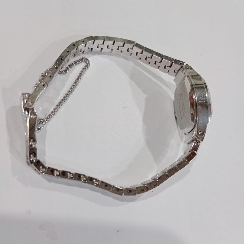 K&ouml;nnte beinhalten: Eine silberfarbene Armbanduhr mit rechteckigem Zifferblatt und einem Metallarmband mit Gliedern. Die Uhr hat eine kleine Kette zur Sicherung des Armbands. Die Uhr wird auf einer wei&szlig;en Oberfl&auml;che pr&auml;sentiert.