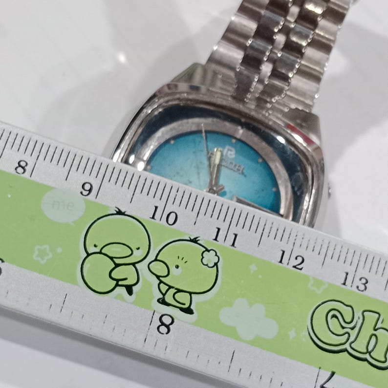 Puede incluir: Un reloj de pulsera plateado con una esfera redonda de color azul brillante y una correa de eslabones met&aacute;licos. Una regla est&aacute; colocada sobre la esfera del reloj, mostrando la medida desde la marca de 8 cm hasta m&aacute;s all&aacute; de los 12 cm.