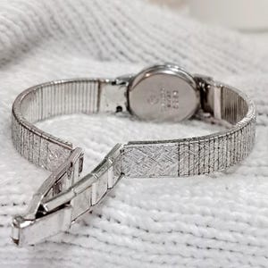 Op de afbeelding: Een zilverkleurig vintage polshorloge met een open sluiting. Het horloge heeft een ronde wijzerplaat en een flexibele, getextureerde band. De band is gedeeltelijk losgemaakt, waardoor het ingewikkelde ontwerp zichtbaar is. Het horloge staat tegen een witte, getextureerde achtergrond.