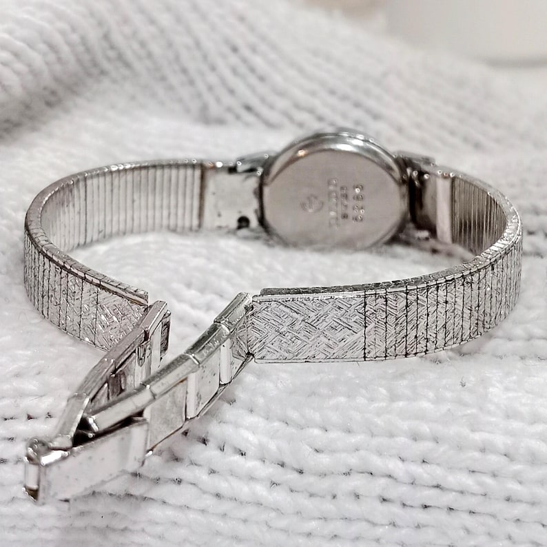 Op de afbeelding: Een zilverkleurig vintage polshorloge met een open sluiting. Het horloge heeft een ronde wijzerplaat en een flexibele, getextureerde band. De band is gedeeltelijk losgemaakt, waardoor het ingewikkelde ontwerp zichtbaar is. Het horloge staat tegen een witte, getextureerde achtergrond.