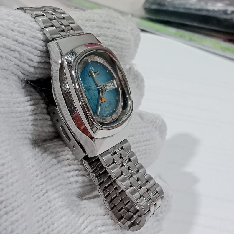 Puede incluir: Un reloj de pulsera autom&aacute;tico Ricoh de tono plateado con una esfera azul turquesa vibrante y una ventana de d&iacute;a y fecha en la posici&oacute;n de las tres. El reloj incluye una caja de acero inoxidable pulido en forma de coj&iacute;n y una correa de eslabones.