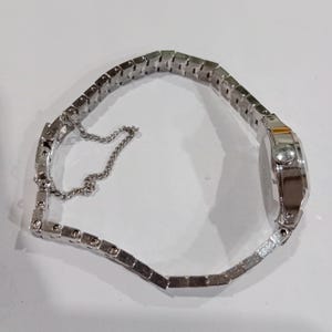 K&ouml;nnte beinhalten: Eine silberfarbene Armbanduhr mit rechteckigem Zifferblatt und einem Metallarmband mit Gliedern. Die Uhr hat ein kleines, rundes Zifferblatt und eine filigrane Kette zur Sicherung des Armbands. Die Uhr liegt auf einer wei&szlig;en Oberfl&auml;che.