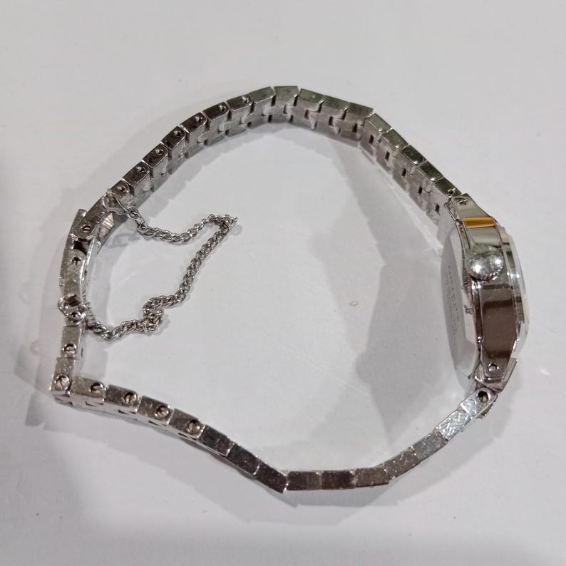 K&ouml;nnte beinhalten: Eine silberfarbene Armbanduhr mit rechteckigem Zifferblatt und einem Metallarmband mit Gliedern. Die Uhr hat ein kleines, rundes Zifferblatt und eine filigrane Kette zur Sicherung des Armbands. Die Uhr liegt auf einer wei&szlig;en Oberfl&auml;che.
