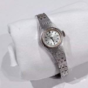 May include: A silver-toned Seiko wristwatch featuring a round dial with thin black hour markers and hands. The watch has a textured metal link bracelet and a circular case with a crown on the right side. The dial displays the Seiko logo and 21 jewels.