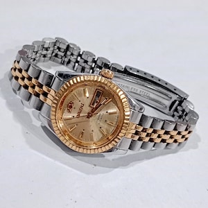 Könnte beinhalten: Eine zweifarbige Orient-Armbanduhr mit goldfarbenem Zifferblatt, Strichindizes, geriffelter Lünette und einem Edelstahl-Gliederarmband mit goldfarbenen Mittelgliedern. Das Zifferblatt zeigt das Orient-Logo sowie Crystal und 21 Jewels.