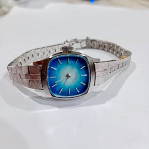 May include: A vintage silver wristwatch with a square face and a blue gradient dial. The watch has a silver strap with a textured pattern. The watch face features silver hour markers and hands.