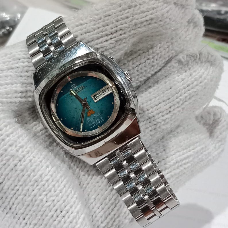 Puede incluir: Un reloj de pulsera autom&aacute;tico Ricoh Crystal plateado con brazalete de eslabones met&aacute;licos. El reloj presenta una esfera redonda de color verde azulado con una ventana de d&iacute;a y fecha a las tres, mostrando 5 FRI y 21 Jewels Automatic.