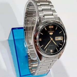May include: A silver-toned Seiko 5 automatic watch with a black dial. The watch features gold-colored hands and markers, and a day-date display. The watchband is made of silver-toned metal links.