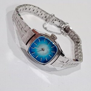 K&ouml;nnte beinhalten: Eine silberfarbene Armbanduhr mit quadratischem Zifferblatt und blau-wei&szlig;em Farbverlauf. Die Uhr hat ein strukturiertes Armband und eine kleine Krone. Das Armband besteht aus kleinen, rechteckigen Gliedern.