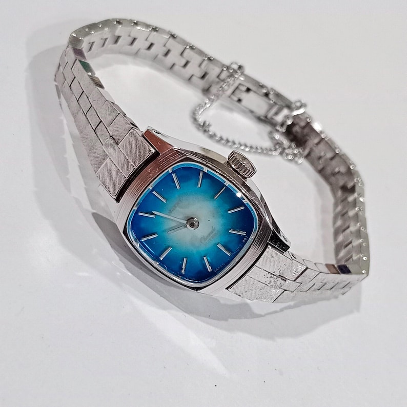 K&ouml;nnte beinhalten: Eine silberfarbene Armbanduhr mit quadratischem Zifferblatt und blau-wei&szlig;em Farbverlauf. Die Uhr hat ein strukturiertes Armband und eine kleine Krone. Das Armband besteht aus kleinen, rechteckigen Gliedern.