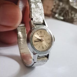 Op de afbeelding: Een vintage Rado polshorloge met een zilverkleurige, getextureerde band en een ronde, goudkleurige wijzerplaat. De wijzerplaat heeft zwarte wijzers en het woord "RADO". Een klassiek horloge.