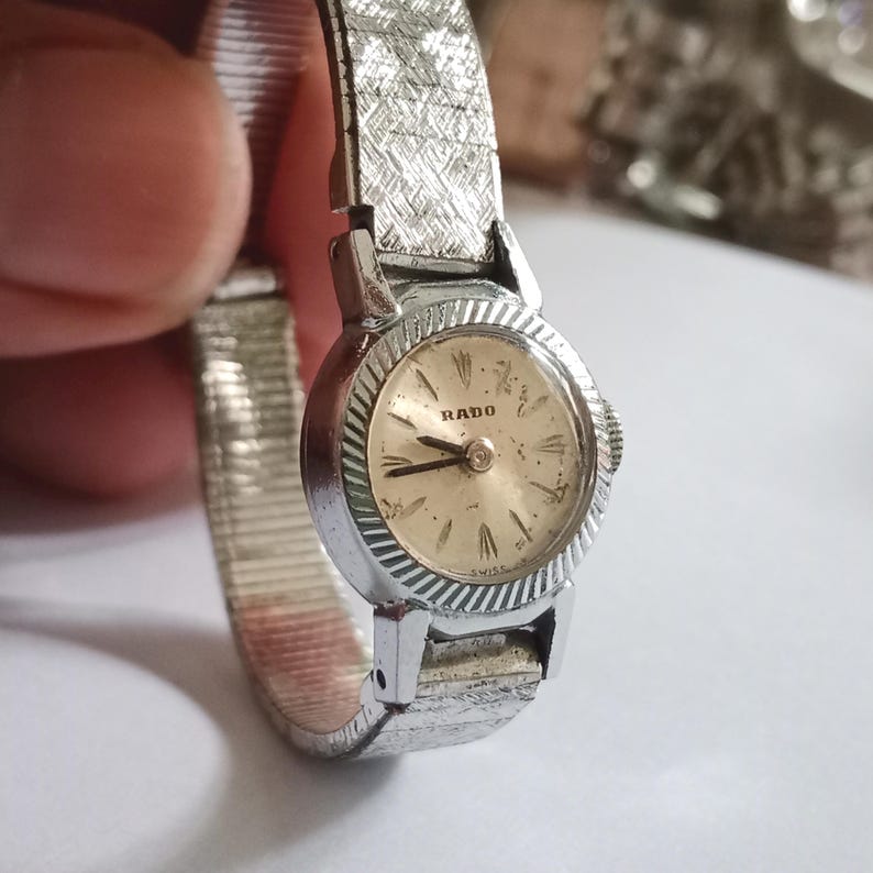 Op de afbeelding: Een vintage Rado polshorloge met een zilverkleurige, getextureerde band en een ronde, goudkleurige wijzerplaat. De wijzerplaat heeft zwarte wijzers en het woord "RADO". Een klassiek horloge.