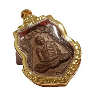 May include: A gold-colored pendant with a detailed relief of a seated figure, possibly a monk or deity, encased in a clear, protective cover. The pendant has an ornate, decorative border and a loop for hanging. The overall design suggests a religious or spiritual item.