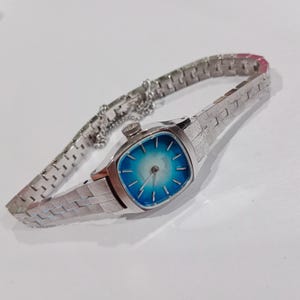 K&ouml;nnte beinhalten: Eine Vintage-Armbanduhr in Silber mit quadratischem Zifferblatt und blauem Farbverlauf. Die Uhr hat ein silbernes Armband mit Kettenverl&auml;ngerung. Ein klassisches Accessoire.