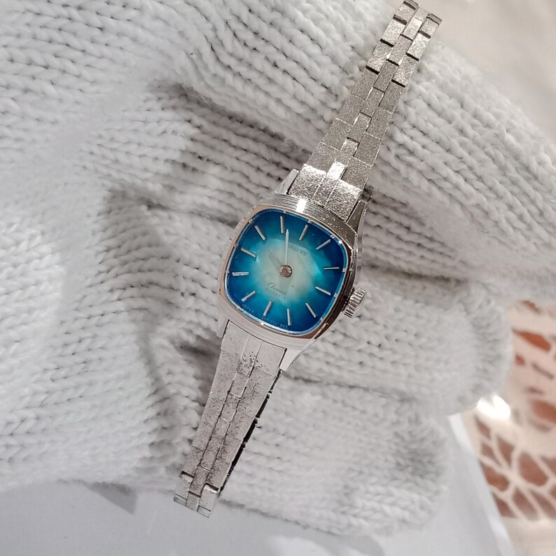 K&ouml;nnte beinhalten: Eine Vintage-Armbanduhr aus Silber mit quadratischem Zifferblatt und blau-t&uuml;rkisfarbenem Farbverlauf. Die Uhr hat ein silbernes Armband mit strukturiertem Design. Die Uhr ist ein klassisches Accessoire.