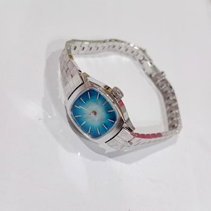 K&ouml;nnte beinhalten: Eine Vintage-Armbanduhr in Silber mit einem quadratischen, hellblauen Zifferblatt. Die Uhr hat ein radiales Design auf dem Zifferblatt und ein silbernes Gliederarmband. Die Uhr ist vor einem wei&szlig;en Hintergrund platziert.