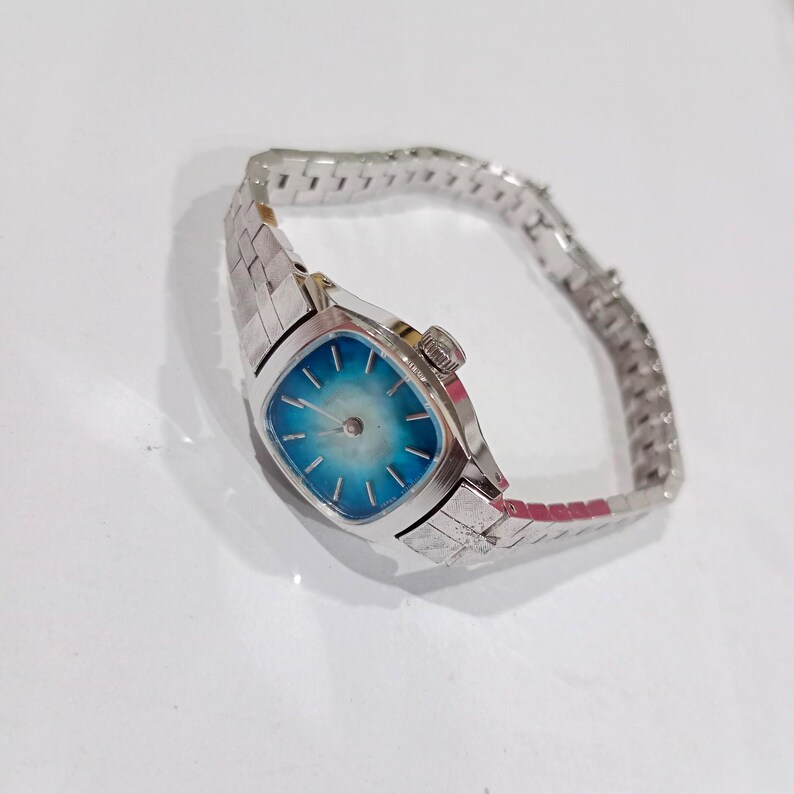 K&ouml;nnte beinhalten: Eine Vintage-Armbanduhr in Silber mit einem quadratischen, hellblauen Zifferblatt. Die Uhr hat ein radiales Design auf dem Zifferblatt und ein silbernes Gliederarmband. Die Uhr ist vor einem wei&szlig;en Hintergrund platziert.