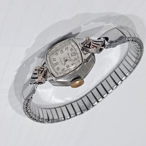 May include: A vintage silver-toned wristwatch with a rectangular face and a stretch band. The watch face is white with black numerals and hands. The watch has gold-toned accents and a gold-toned crown. The watch is a classic timepiece.
