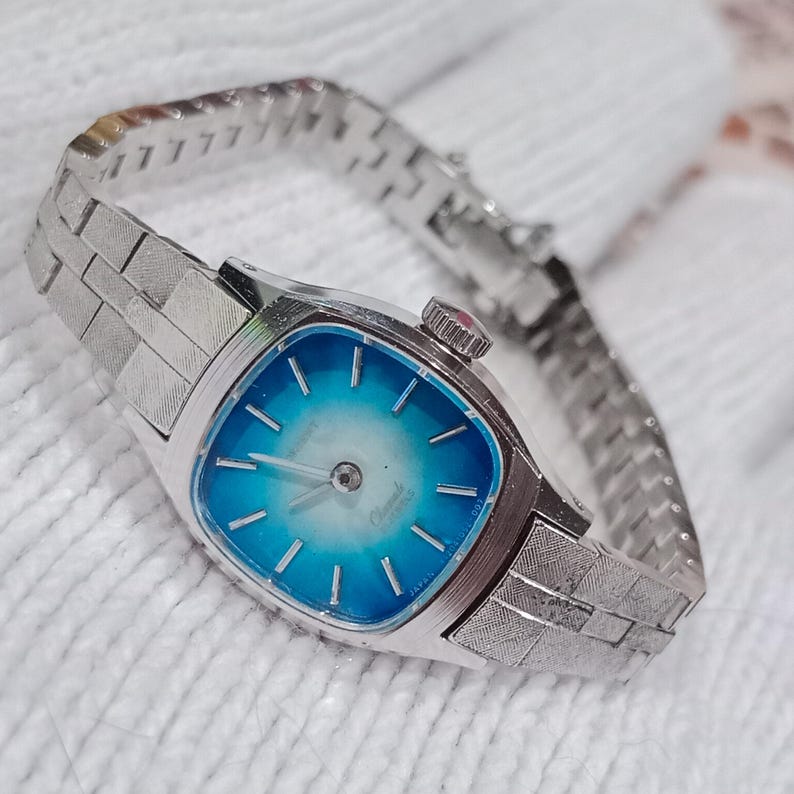 K&ouml;nnte beinhalten: Eine Vintage-Armbanduhr in Silber mit quadratischem Zifferblatt und blauem Farbverlauf. Die Uhr hat ein silbernes Armband mit strukturiertem Design. Auf dem Zifferblatt steht "Chantal" und "Japan".