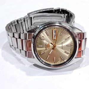 May include: A Seiko 5 automatic wristwatch with a silver-tone stainless steel link bracelet and a round champagne-colored dial. The watch face features silver-tone hour markers, luminous hands, and a day-date window at the three o'clock position.